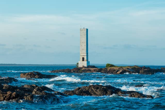 Um alto farol de concreto erguido sobre um ilhote rochoso rodeado pelas ondas azuis do Atlântico próximo a Itacaré, Bahia, Brasil