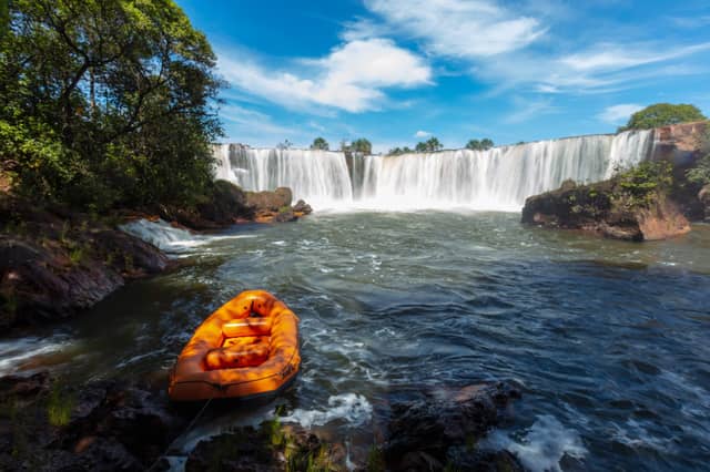 An orange inflatable raft moored on rocky riverbank in front of a wide curtain waterfall surrounded by Cerrado forest on a Jalapão tour in Brazil