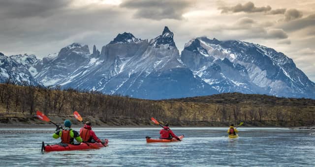 Four kayakers in red and yellow sea kayaks paddling across a glacial lake with the jagged snow-capped peaks of Torres del Paine rising dramatically under a cloudy sky in Chilean Patagonia