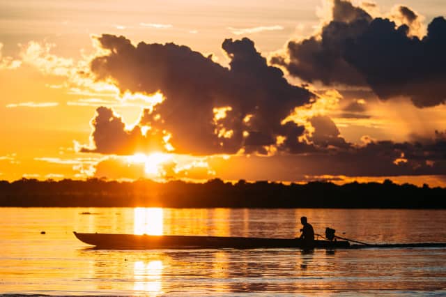 Silhueta de uma pessoa em uma longa canoa de madeira sobre um rio amplo ao pôr do sol, com céu alaranjado intenso e nuvens de tempestade dramáticas sobre o horizonte arborizado perto de Iquitos
