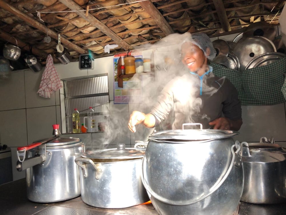 Eine einheimische Frau lächelt, während sie mit großen Aluminiumtöpfen auf einem Herd eine traditionelle hausgemachte Mahlzeit kocht.