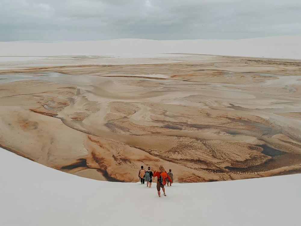 Um grupo de turistas caminha pelo leito seco e marrom de uma lagoa, cercado por grandes dunas de areia branca durante a estação seca.