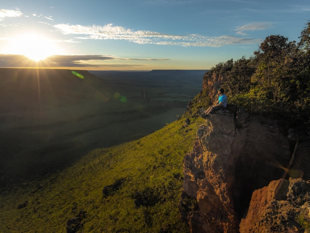 Een reiziger zit op de rand van een klif en geniet van de zonsondergang over het uitgestrekte savannelandschap van het Jalapão State Park.