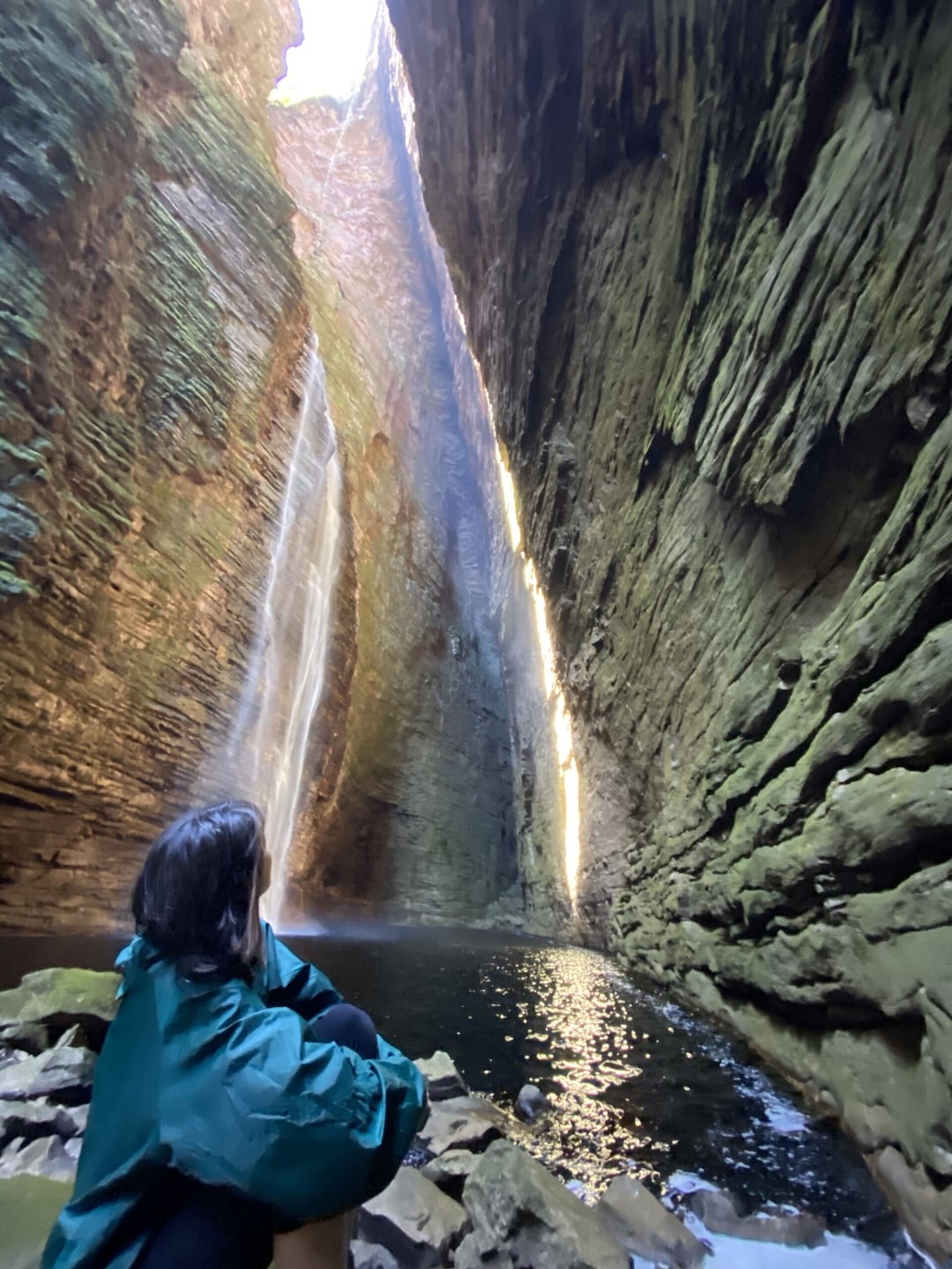 Um trilheiro de jaqueta verde admira uma cachoeira imponente dentro de um cânion íngreme durante a estação chuvosa na Chapada Diamantina.