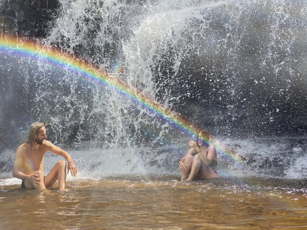 Dois viajantes sentados na parte rasa da base de uma cachoeira com o reflexo de um arco-íris.