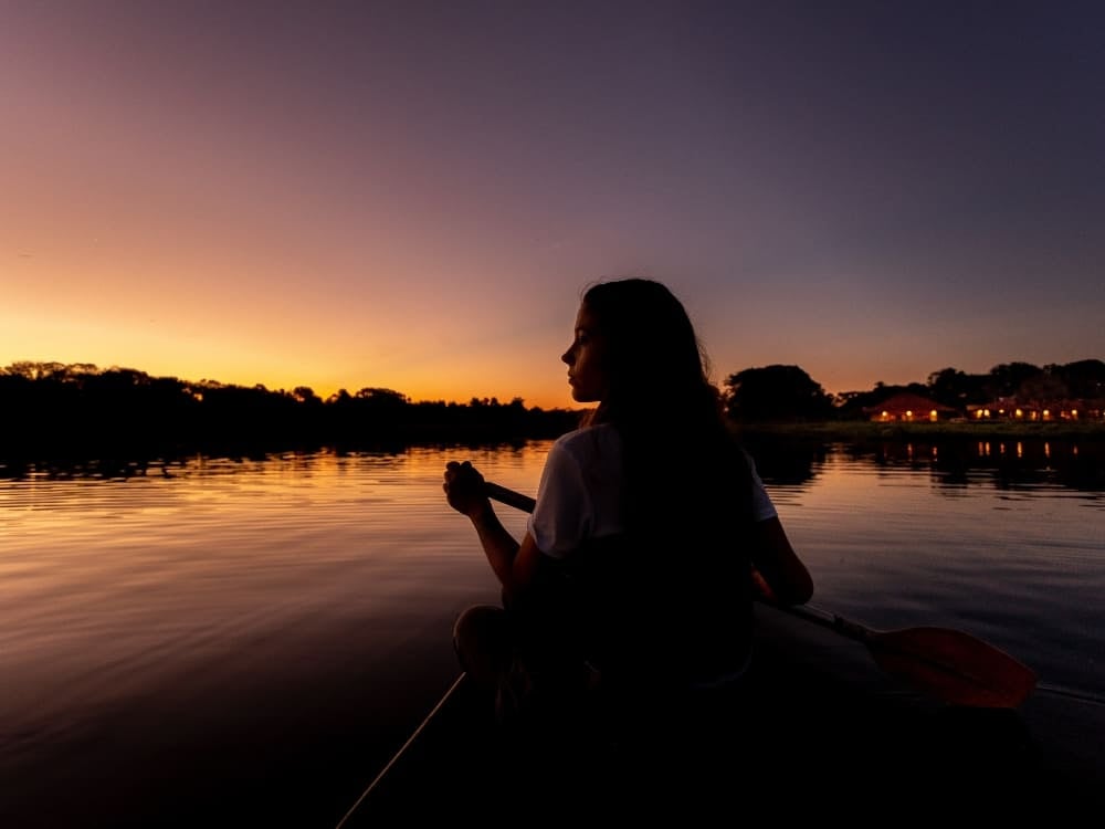 Uma mulher remando uma canoa em um rio calmo ao pôr do sol, destacando a natureza pacífica de uma viagem ao Pantanal.
