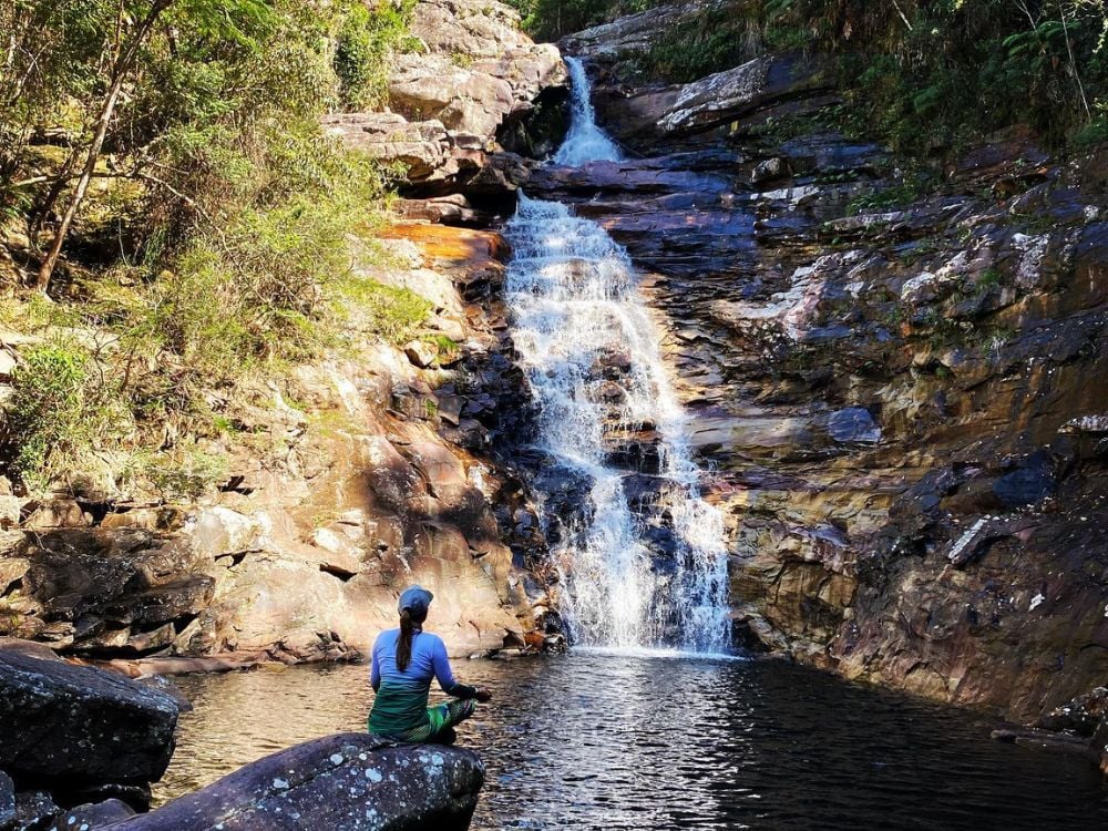 Uma mulher sentada pacificamente em uma rocha à beira da piscina natural na base da Cachoeira dos Funis, com 30 metros de altura.