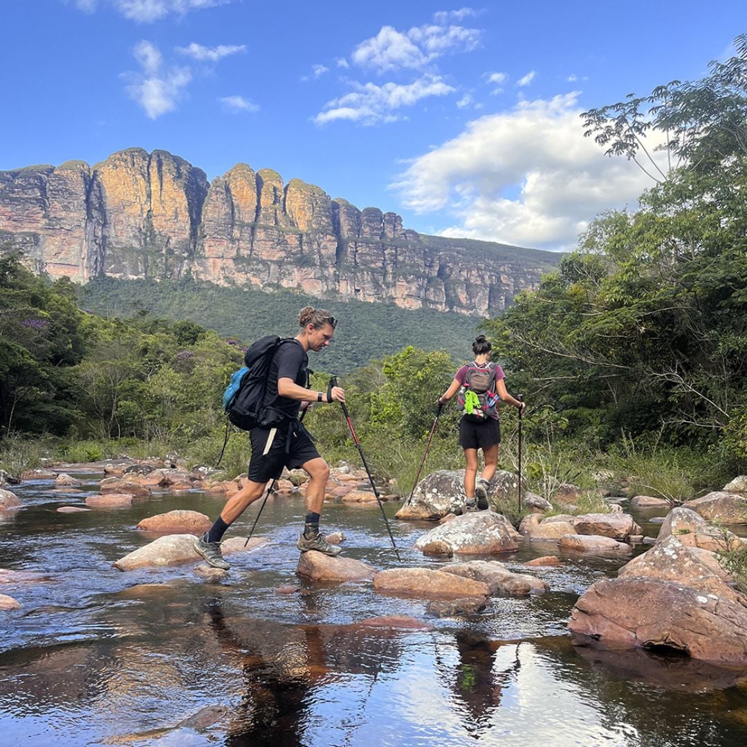 Dois trilheiros usando bastões de caminhada para cruzar um rio raso e pedregoso com um grande pico de montanha erguendo-se ao fundo.
