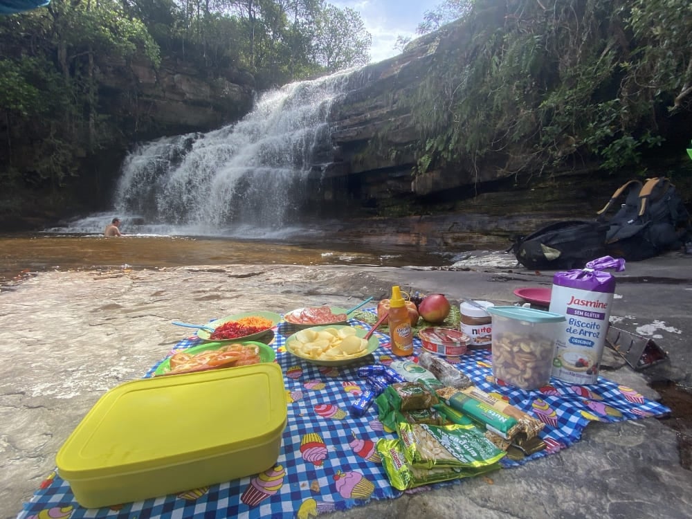 Um piquenique colorido com frutas, lanches e sanduíches dispostos sobre uma toalha ao lado de uma pequena cascata.