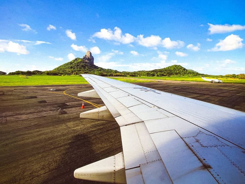 Vista da janela de um avião mostrando a asa e o famoso Morro do Pico, enquanto um voo chega a Fernando de Noronha.