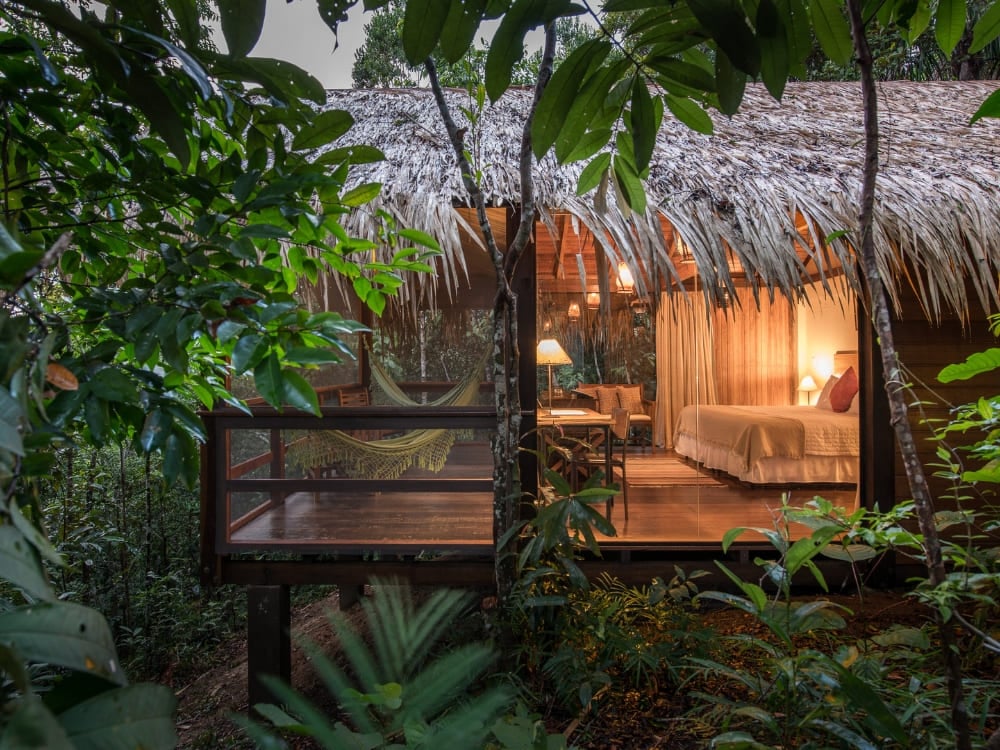 A luxury thatched-roof bungalow with warm interior lighting sits nestled among lush green trees at the Anavilhanas Jungle Lodge.