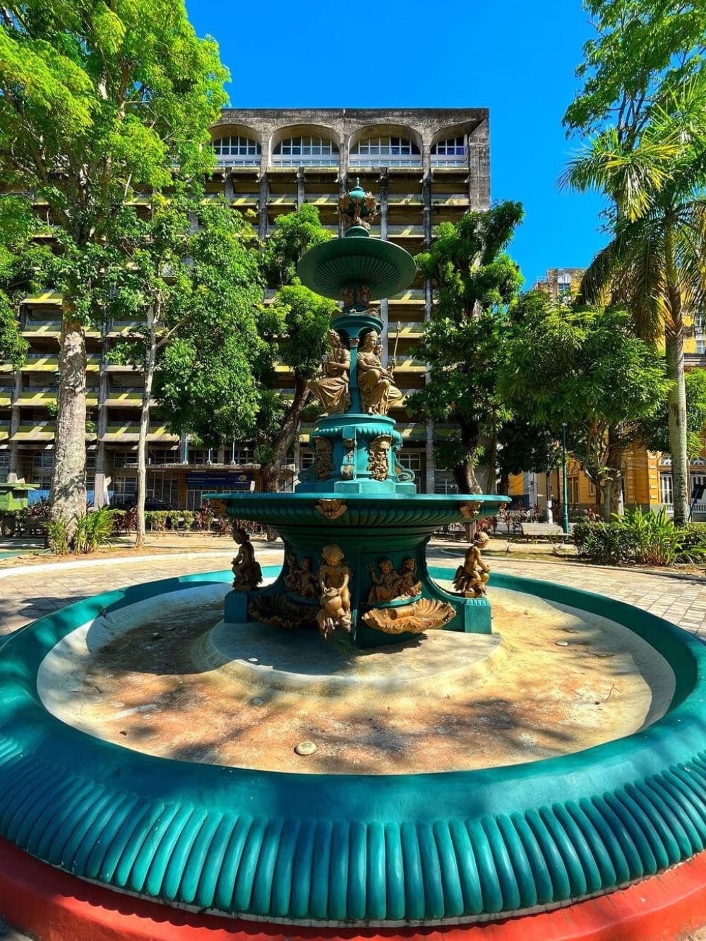Green iron fountain located in a public square within the historic center of Manaus surrounded by trees.