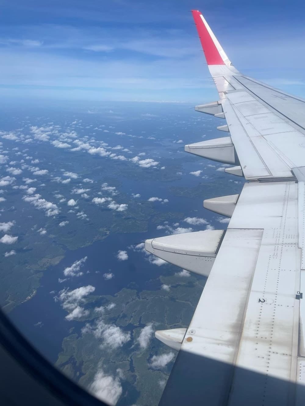 View from an airplane window looking out over the wing towards the Amazon clouds and landscape below.