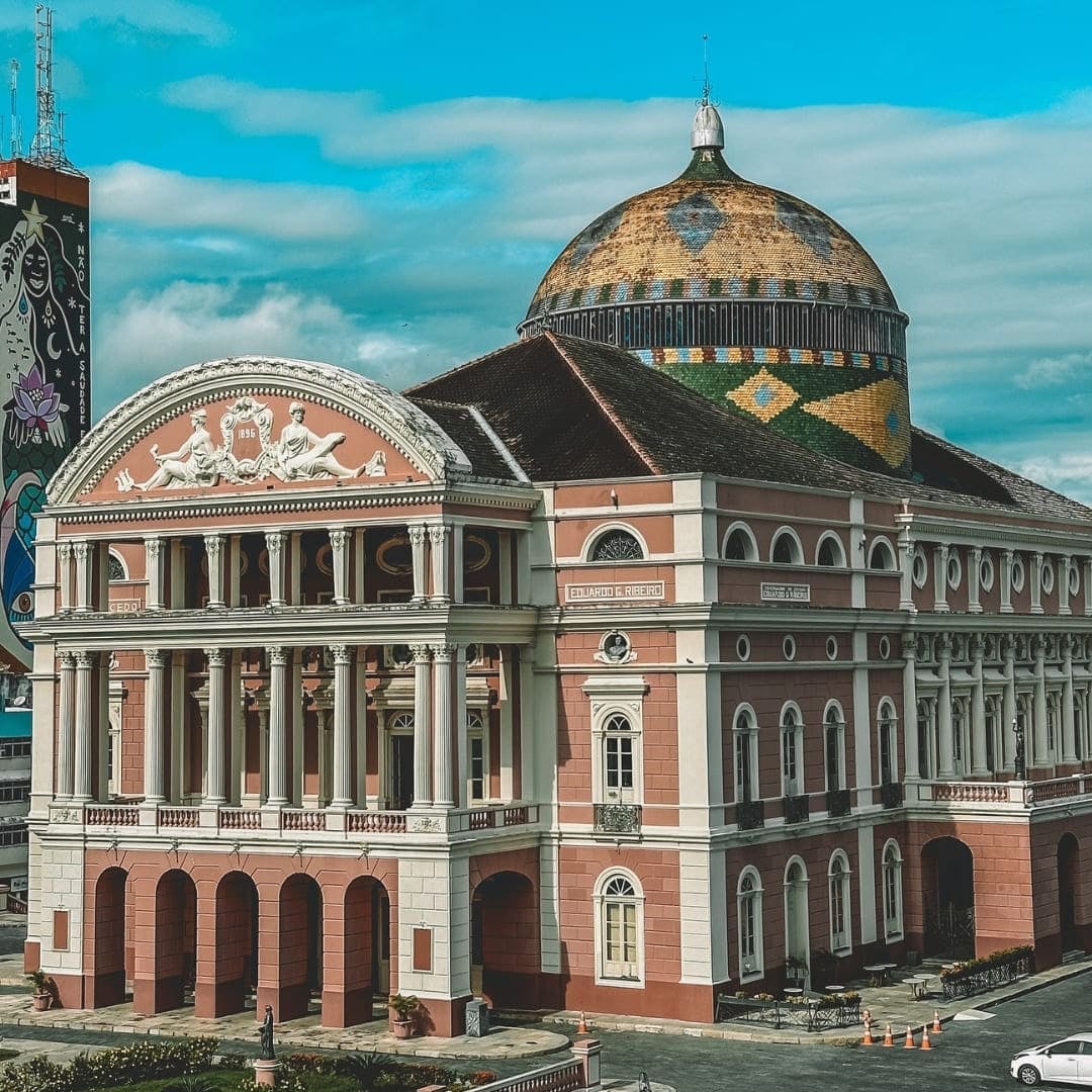 The pink neoclassical facade and colorful tiled dome of the Amazon Theatre in Manaus under a blue sky.