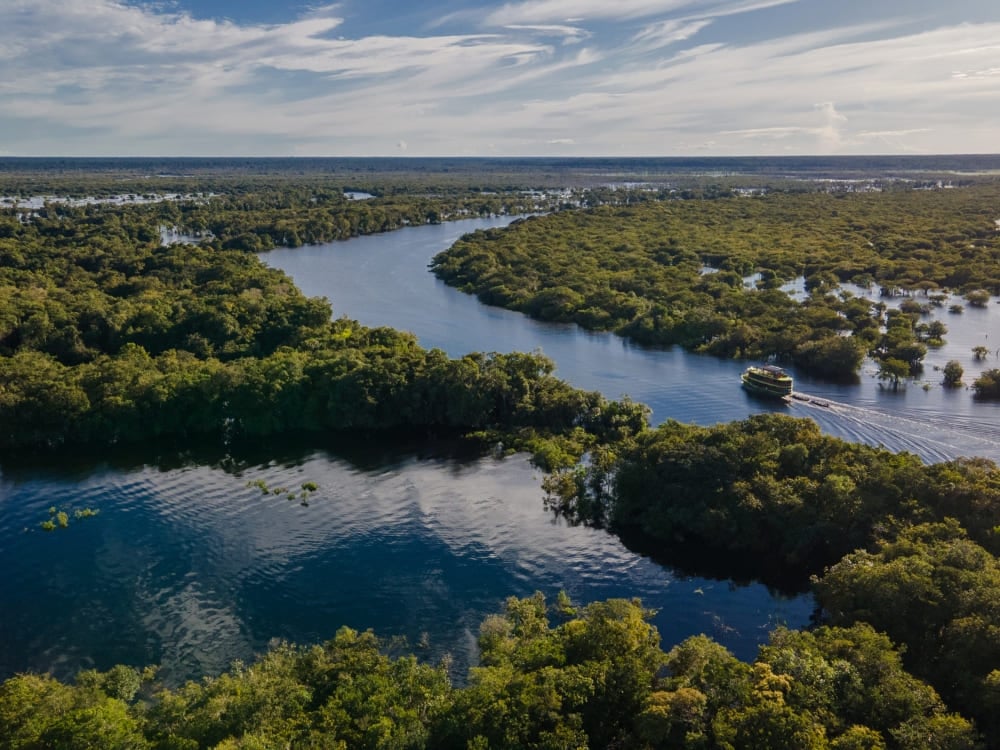 Vista aérea de um barco regional navegando pela bacia do rio Jaú, cercado pela imensa floresta verdejante da Amazônia.