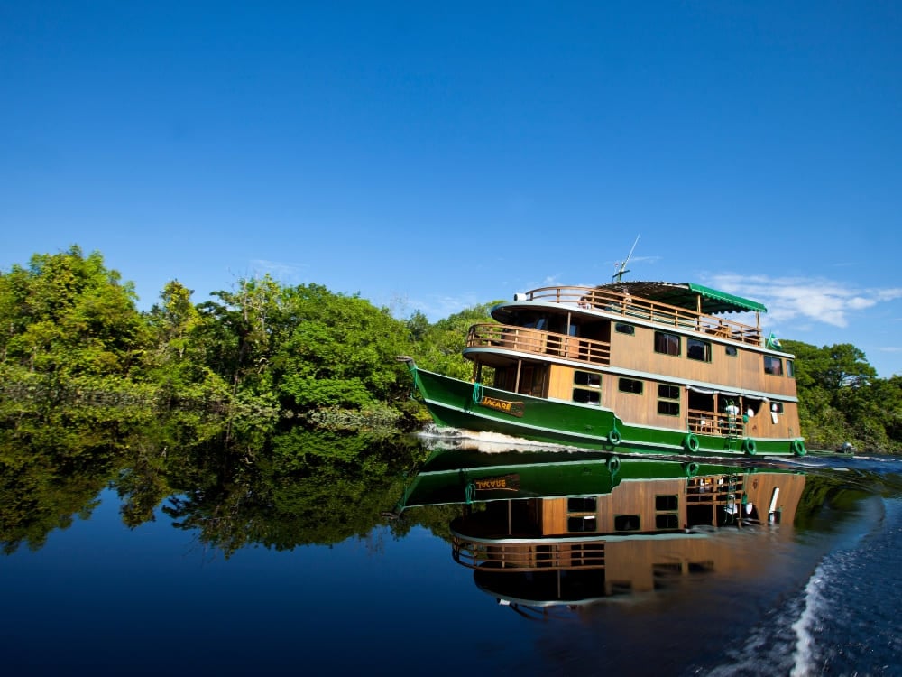 Um barco regional de madeira rústico com três andares navega suavemente por um rio escuro da Amazônia sob um céu azul.