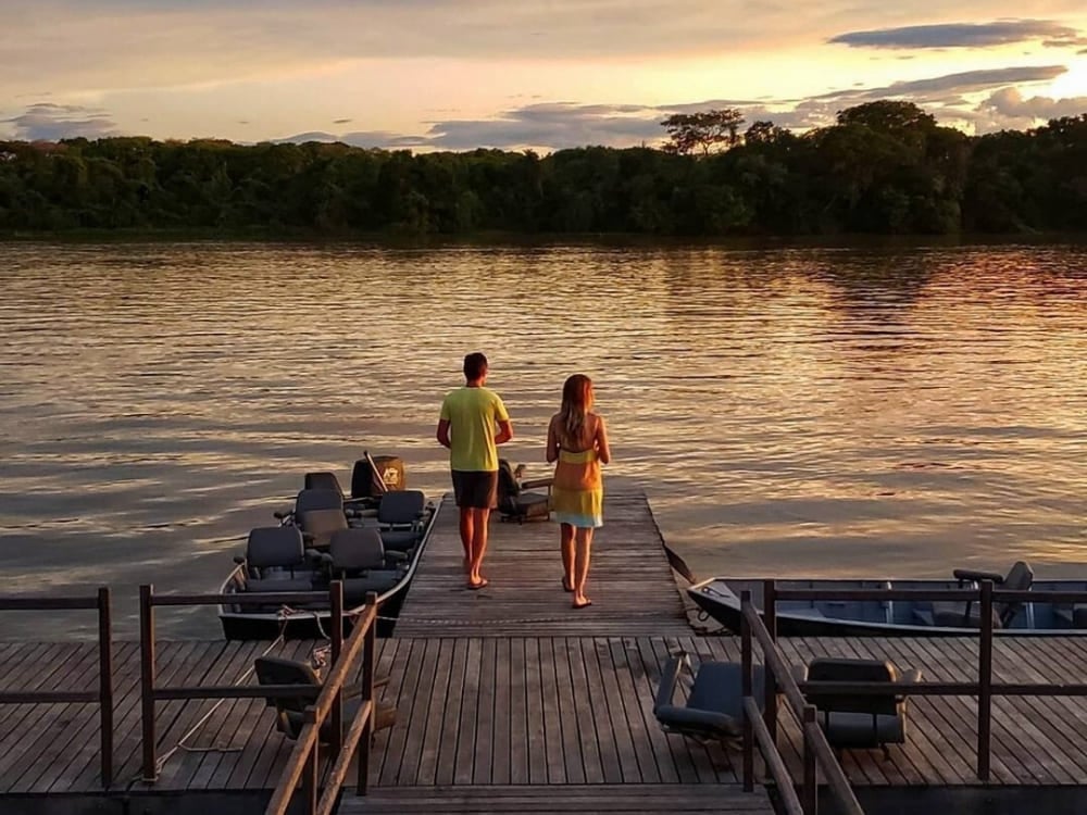 Un couple marche sur un ponton en bois vers des bateaux de safari lors d'un magnifique coucher de soleil sur une rivière du Pantanal.