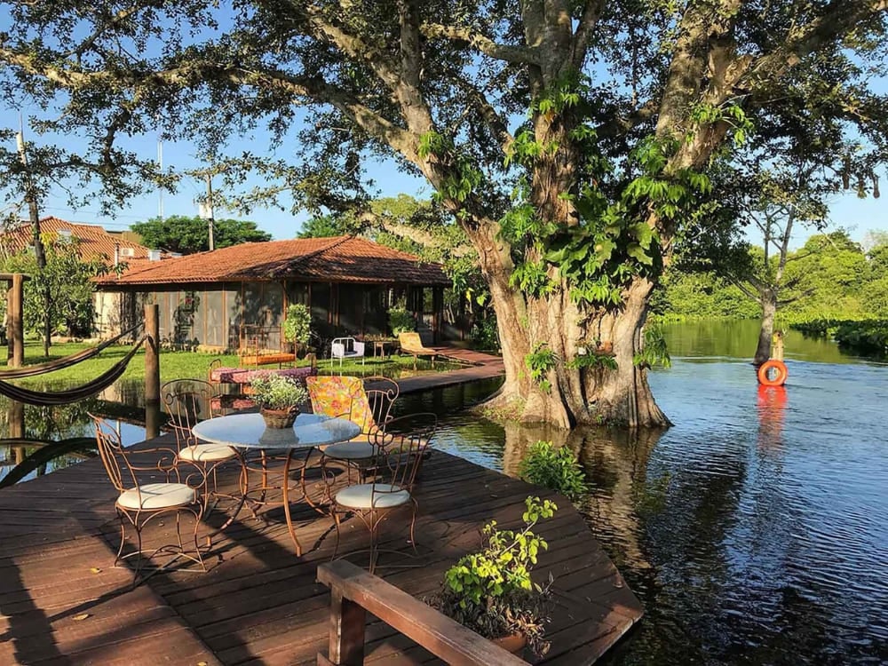 Une terrasse en bois avec une table et des chaises est située à côté d'un lac tranquille et d'un arbre massif à l'auberge écologique Refúgio da Ilha.