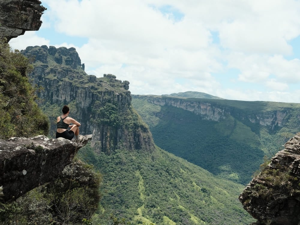 Iemand zit op de rand van een rotsklif en kijkt uit over de diepe, groene boskloven van Chapada Diamantina.