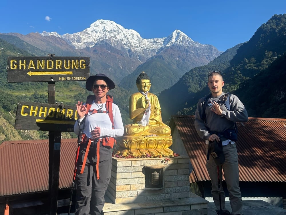 A woman and a young man with hiking backpacks smile next to a golden Buddha statue, with the majestic snow-capped Himalayan mountains in the background.