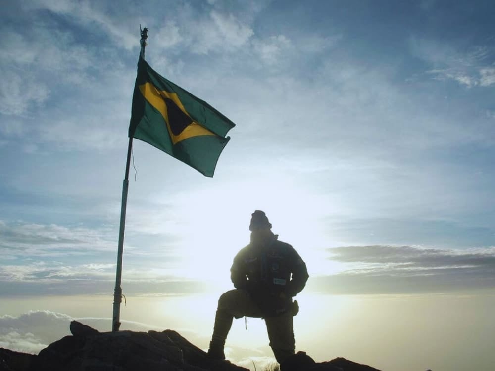 Silhueta de um explorador ajoelhado perto de uma bandeira brasileira tremulando no cume do Pico da Neblina ao nascer do sol.