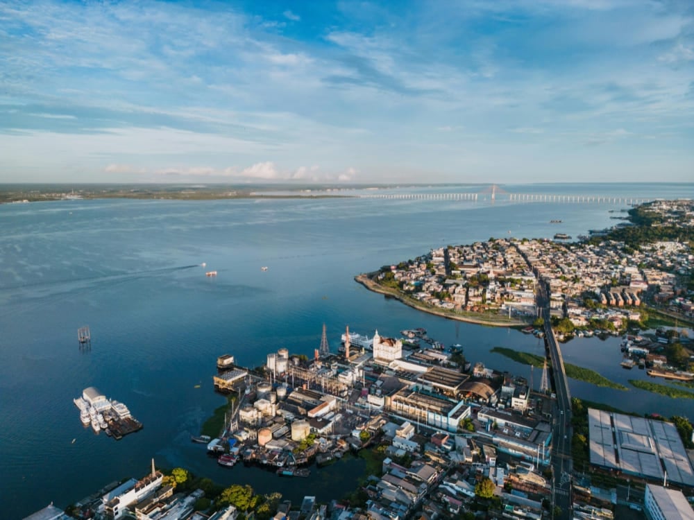 Vista aérea de Manaus, Amazonas, mostrando o porto da cidade, bairros residenciais e o extenso rio com uma longa ponte ao fundo.