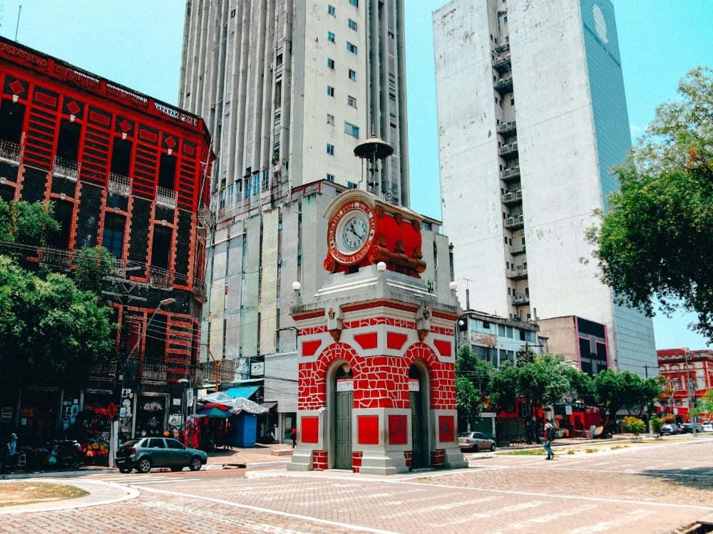 Histórica torre do relógio em pedra e tijolos avermelhados, erguida em uma praça com o contraste de modernos prédios altos em Manaus.