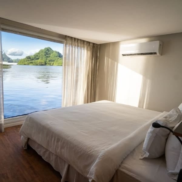 A bright Amazon river cruise cabin with a large bed and a picture window overlooking the river and a forested island.