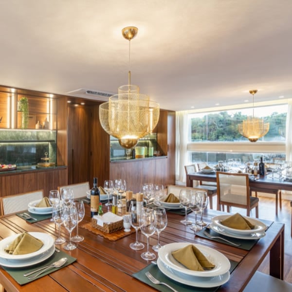 An elegant dining room aboard an Amazon river cruise, a wooden table set with wine glasses under gold chandeliers and windows onto the river.