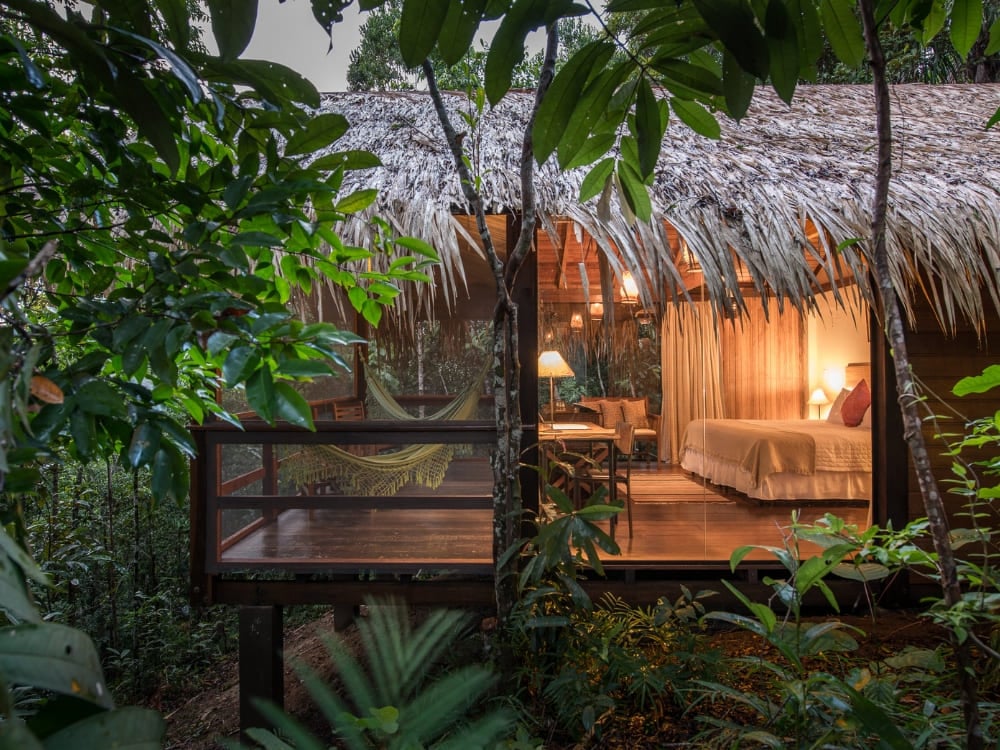 Um bangalô de pousada de luxo na Amazônia, com teto de palha e paredes de vidro iluminadas por luz quente ao anoitecer, com uma rede no deck em meio à floresta.
