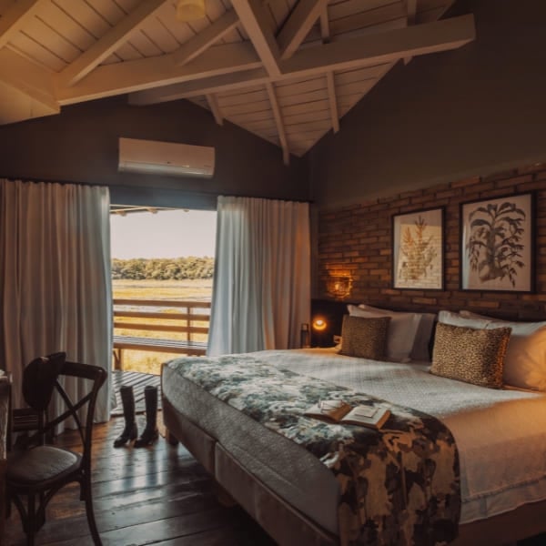 A rustic-chic Pantanal lodge suite with a large bed, a white vaulted beam ceiling and an exposed brick wall, opening through a wide window onto a golden floodplain view.