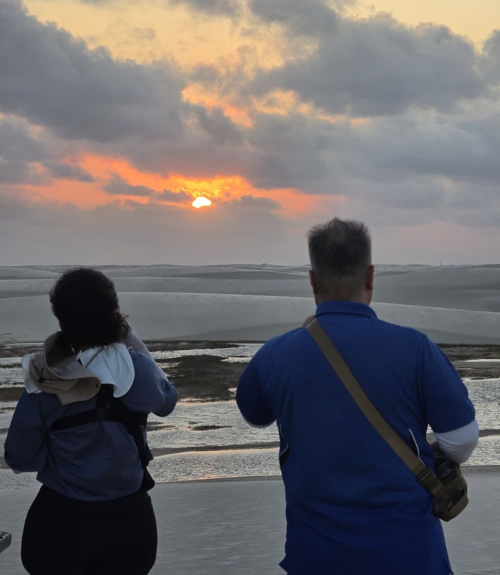 Due viaggiatori guardano il sole che filtra tra le nuvole sopra le dune e le lagune dei Lençóis Maranhenses