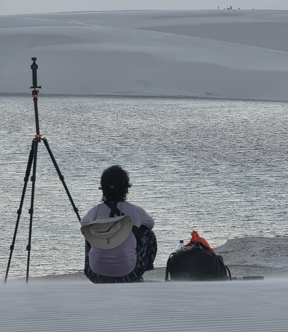 Una persona seduta su una duna accanto a un treppiede, davanti a una laguna nei Lençóis Maranhenses