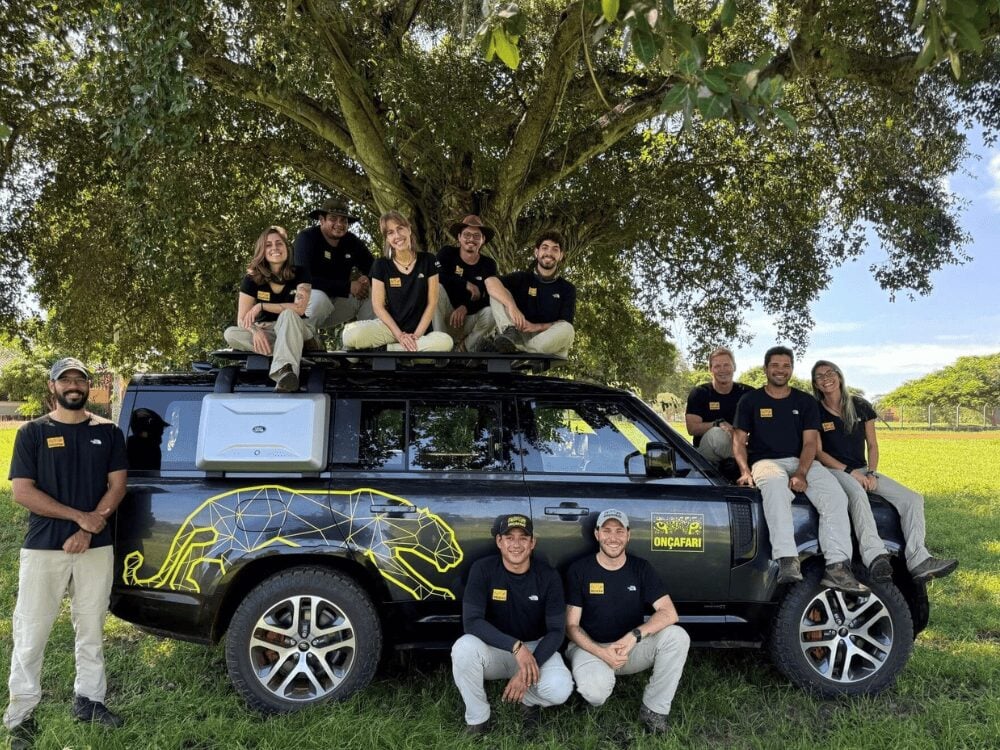 Onçafari team in branded shirts gathered around a Land Rover Defender with a yellow jaguar painted on the side
