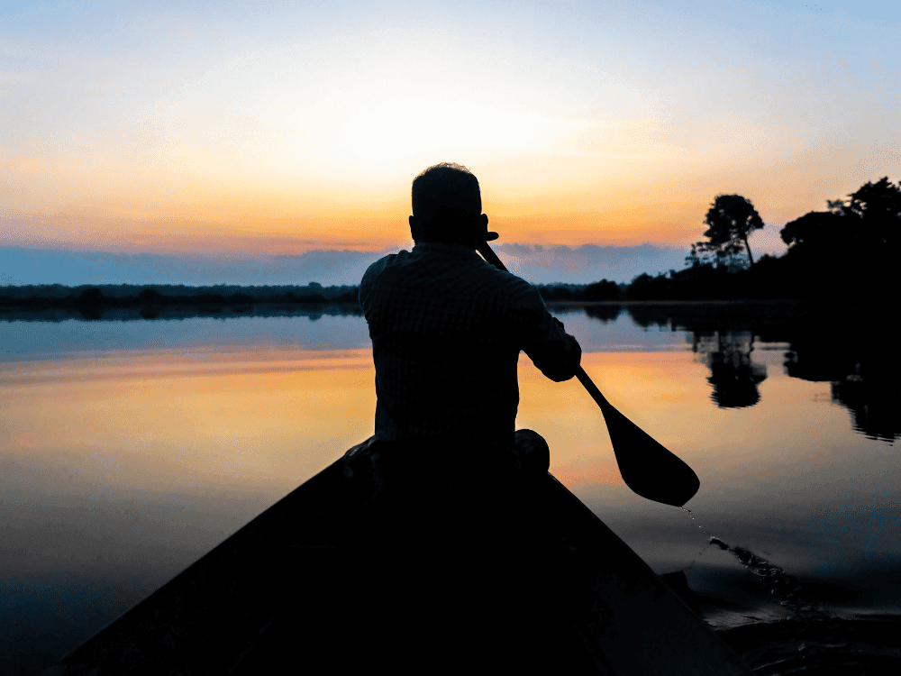 Silhueta de um homem remando uma canoa num rio calmo ao pôr do sol, um jeito mais tranquilo de explorar a Amazônia
