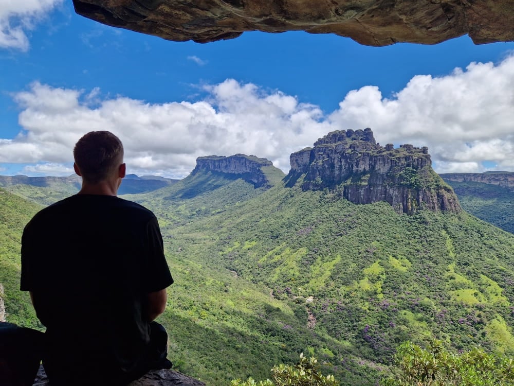 Um homem sentado sob uma saliência rochosa observando um vasto vale verde e montanhas de topo plano na Chapada Diamantina. Um homem sentado sob uma saliência rochosa observando um vasto vale verde e montanhas de topo plano na Chapada Diamantina.