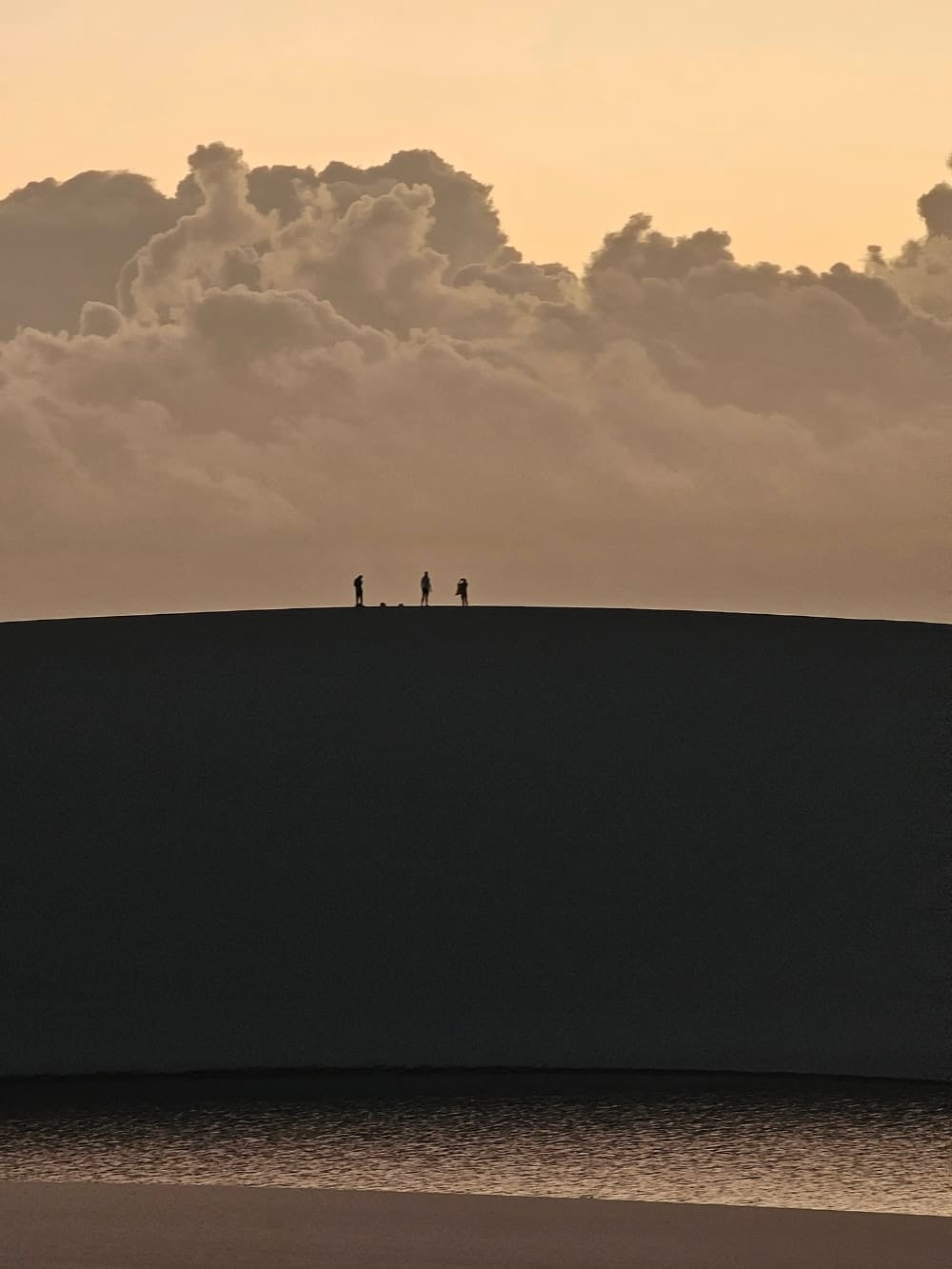 Silhuetas de três pessoas caminhando no topo de uma duna de areia nos Lençóis Maranhenses durante o nascer do sol, com nuvens ao fundo. Silhuetas de três pessoas caminhando no topo de uma duna de areia nos Lençóis Maranhenses durante o nascer do sol, com nuvens ao fundo.