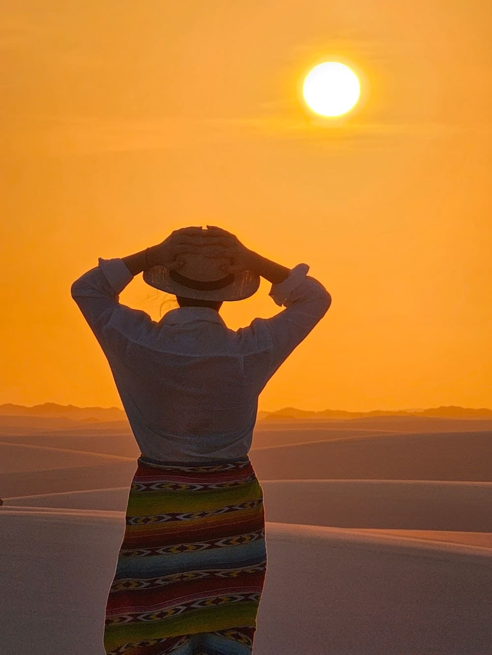 Mulher de costas, usando um chapéu e uma saia colorida, contemplando o pôr do sol dourado sobre a vasta extensão de dunas no parque nacional. Mulher de costas, usando um chapéu e uma saia colorida, contemplando o pôr do sol dourado sobre a vasta extensão de dunas no parque nacional.