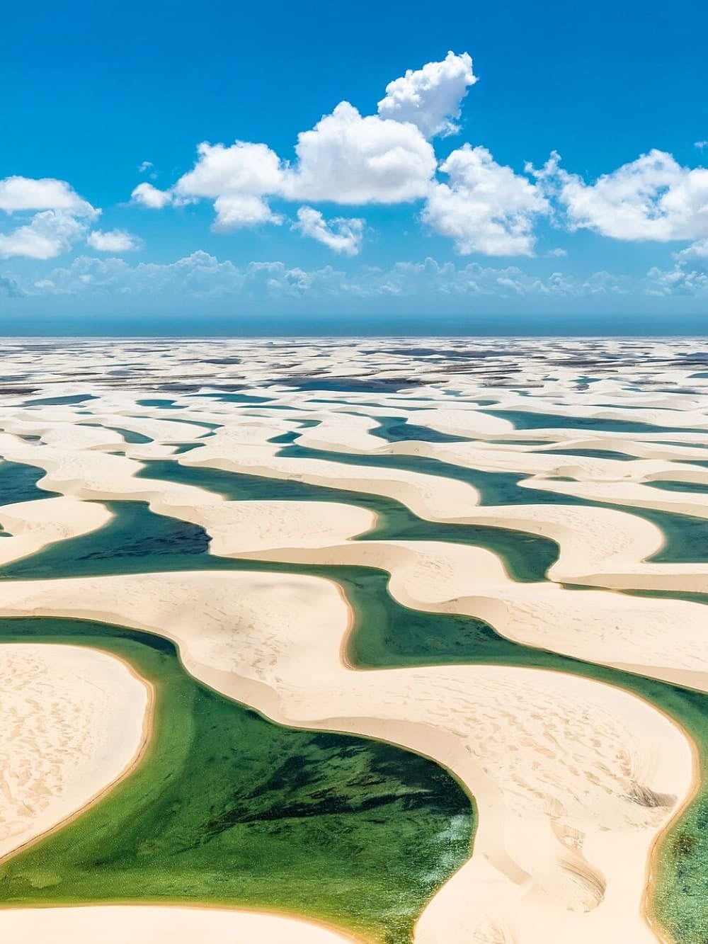 Vista aérea de dunas de areia branca e lagoas de água doce se estendendo pela paisagem dos Lençóis Maranhenses sob um céu azul. Vista aérea de dunas de areia branca e lagoas de água doce se estendendo pela paisagem dos Lençóis Maranhenses sob um céu azul.