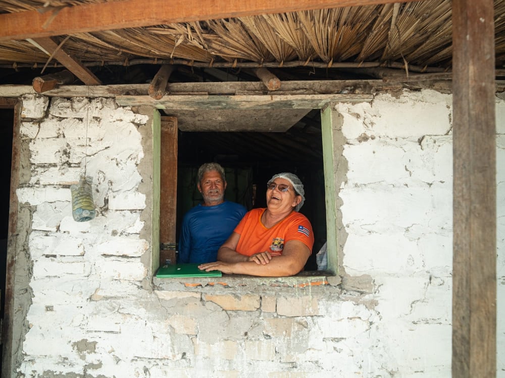 Um homem mais velho sorridente e uma mulher olham pela janela de uma casa rústica, pintada de branco, com telhado de palha nos Lençóis Maranhenses. Um homem mais velho sorridente e uma mulher olham pela janela de uma casa rústica, pintada de branco, com telhado de palha nos Lençóis Maranhenses.