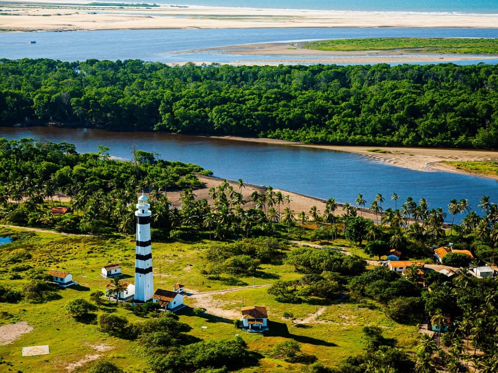 Vista aérea de um farol alto com listras pretas e brancas e uma vegetação verde e exuberante perto das cidades-base para passeios nos Lençóis Maranhenses. Vista aérea de um farol alto com listras pretas e brancas e uma vegetação verde e exuberante perto das cidades-base para passeios nos Lençóis Maranhenses.