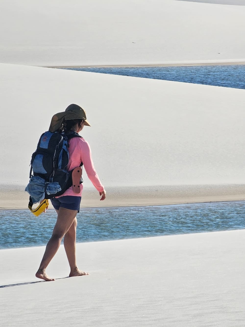 Um mochileiro caminhando descalço pelas dunas de areia branca perto de uma lagoa azul brilhante durante um trekking fisicamente exigente nos Lençóis Maranhenses. Um mochileiro caminhando descalço pelas dunas de areia branca perto de uma lagoa azul brilhante durante um trekking fisicamente exigente nos Lençóis Maranhenses.