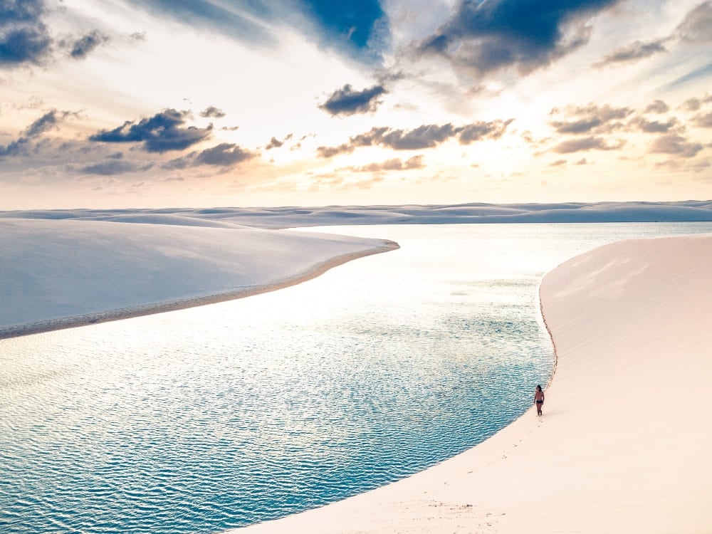 Um viajante caminhando pela beira de uma lagoa azul cristalina cercada por dunas de areia durante a melhor época para visitar os Lençóis Maranhenses. Um viajante caminhando pela beira de uma lagoa azul cristalina cercada por dunas de areia durante a melhor época para visitar os Lençóis Maranhenses.