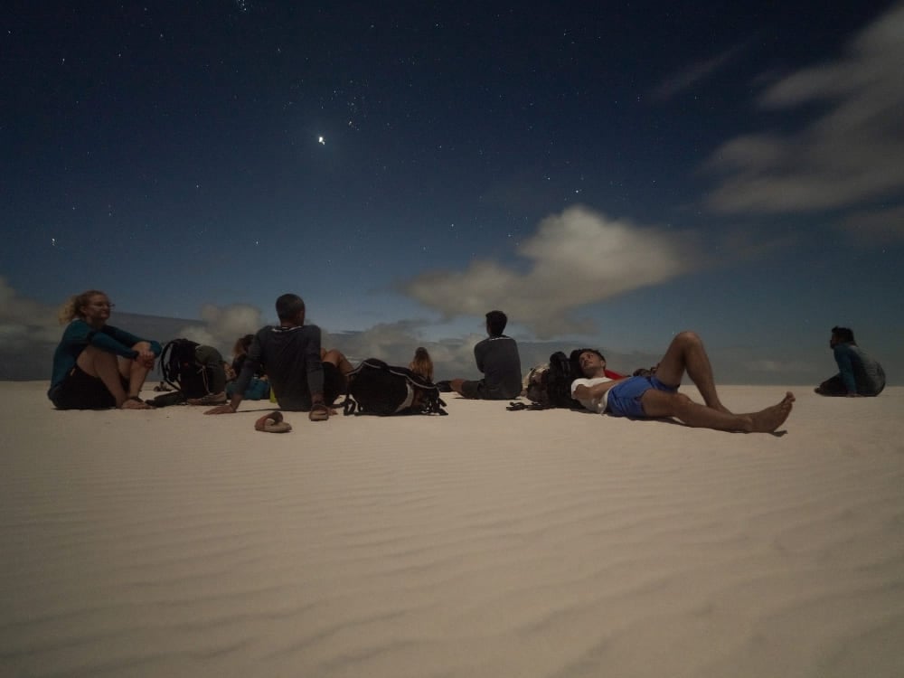 Um grupo de viajantes descansando nas dunas de areia sob um céu noturno estrelado após um dia inteiro explorando os Lençóis Maranhenses. Um grupo de viajantes descansando nas dunas de areia sob um céu noturno estrelado após um dia inteiro explorando os Lençóis Maranhenses.