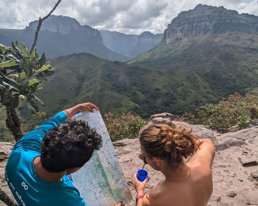 Um guia local do PlanetaEXO vestindo uma camisa azul mostra um mapa de trilhas para uma mulher trilheira, com um vasto vale verde e montanhas ao fundo. Um guia local do PlanetaEXO vestindo uma camisa azul mostra um mapa de trilhas para uma mulher trilheira, com um vasto vale verde e montanhas ao fundo.