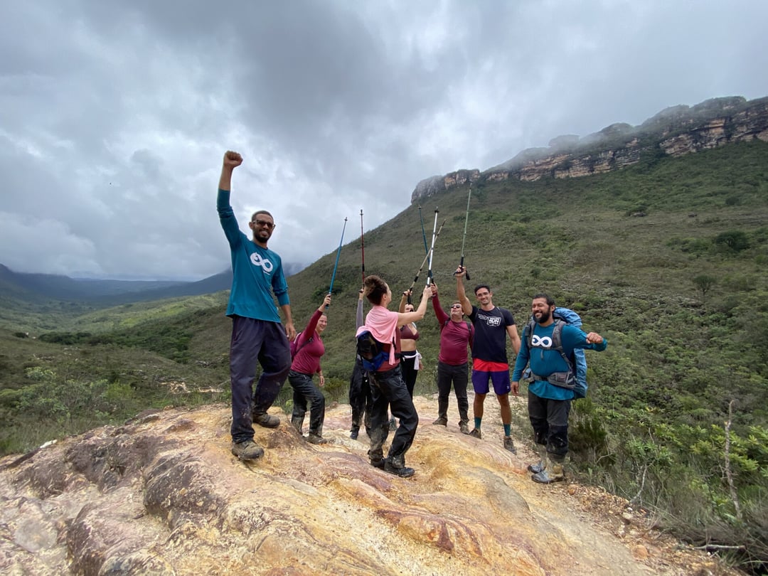 Um grupo de trilheiros e um guia do PlanetaEXO comemoram com os braços erguidos em um pico rochoso, cercados por montanhas verdes sob um céu nublado. Um grupo de trilheiros e um guia do PlanetaEXO comemoram com os braços erguidos em um pico rochoso, cercados por montanhas verdes sob um céu nublado.