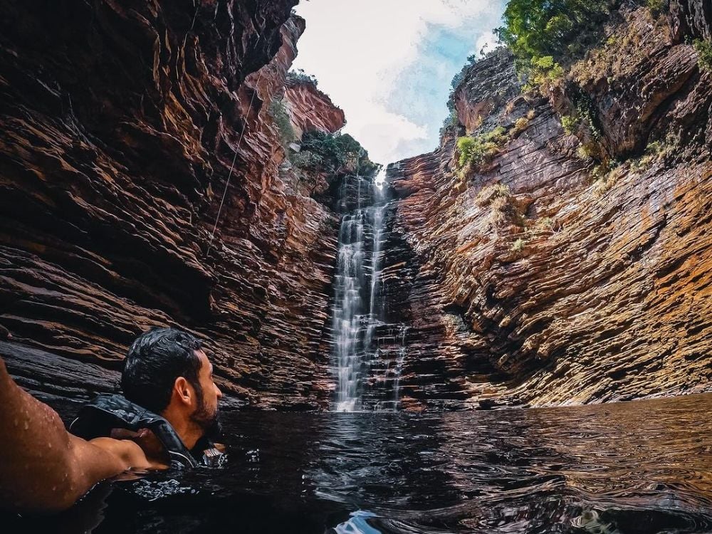 Um visitante nada em um poço escuro sob uma cachoeira cercada por imponentes formações rochosas em camadas na Chapada Diamantina. Um visitante nada em um poço escuro sob uma cachoeira cercada por imponentes formações rochosas em camadas na Chapada Diamantina.