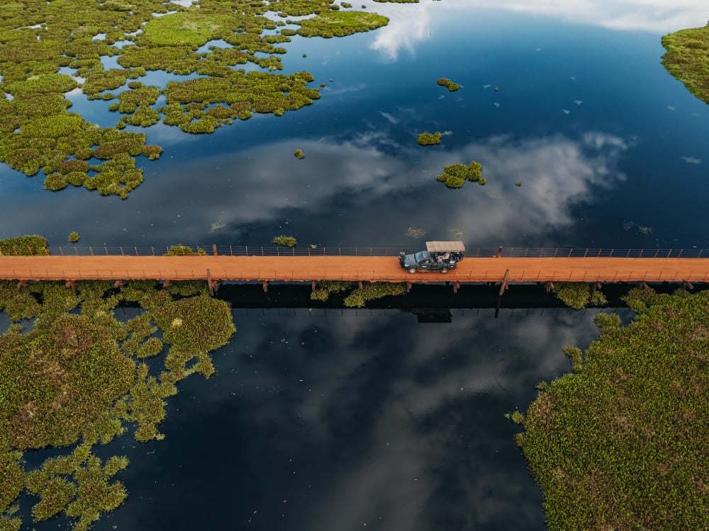 Uma vista aérea de um veículo de safári cruzando uma longa ponte de madeira sobre as áreas alagadas durante um safári no Pantanal no Brasil. Uma vista aérea de um veículo de safári cruzando uma longa ponte de madeira sobre as áreas alagadas durante um safári no Pantanal no Brasil.