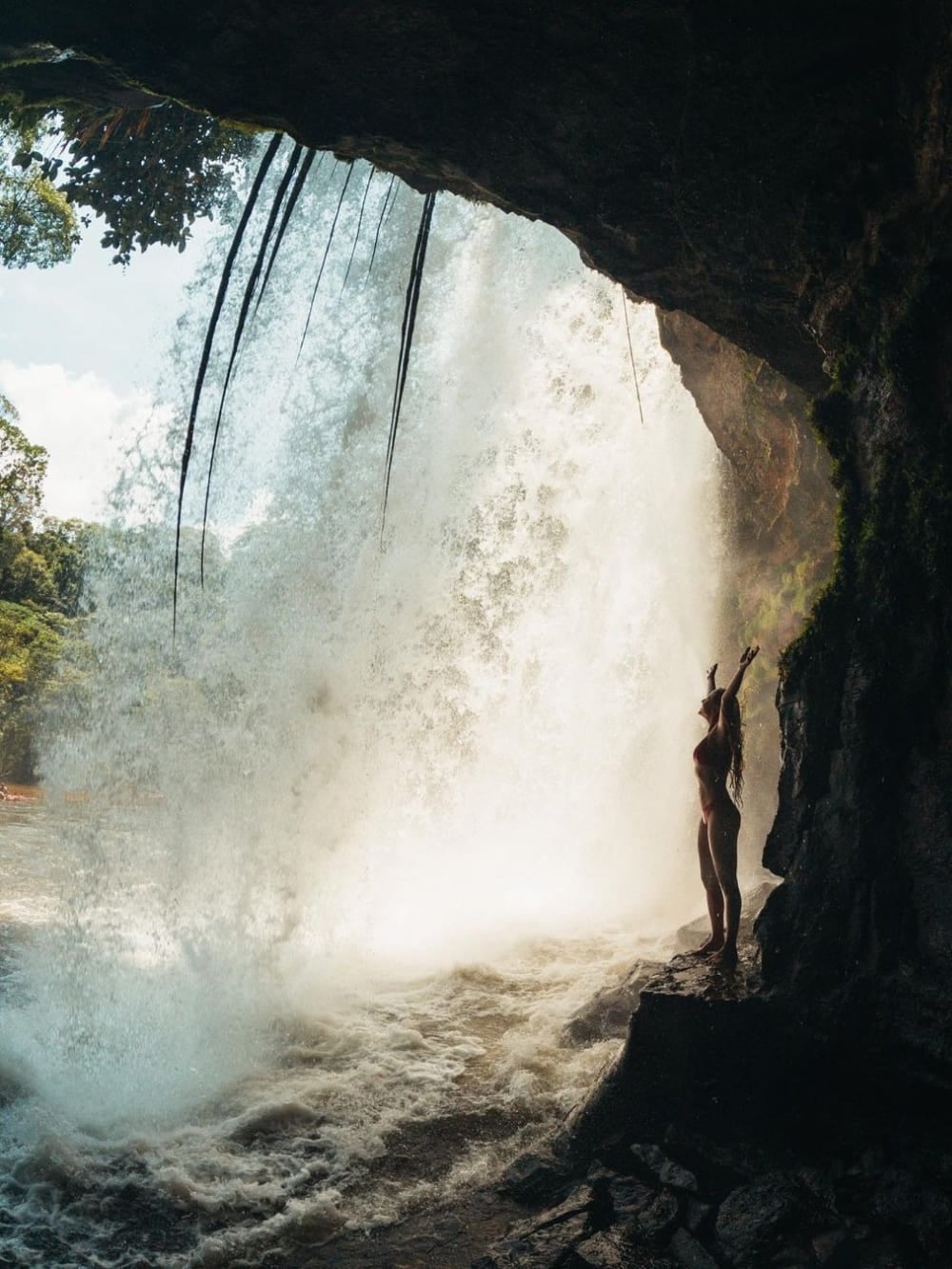 Die Silhouette einer Frau mit offenen Armen, die aus dem dunklen Inneren einer Höhle einen kräftigen Wasserfall betrachtet. Die Silhouette einer Frau mit offenen Armen, die aus dem dunklen Inneren einer Höhle einen kräftigen Wasserfall betrachtet.