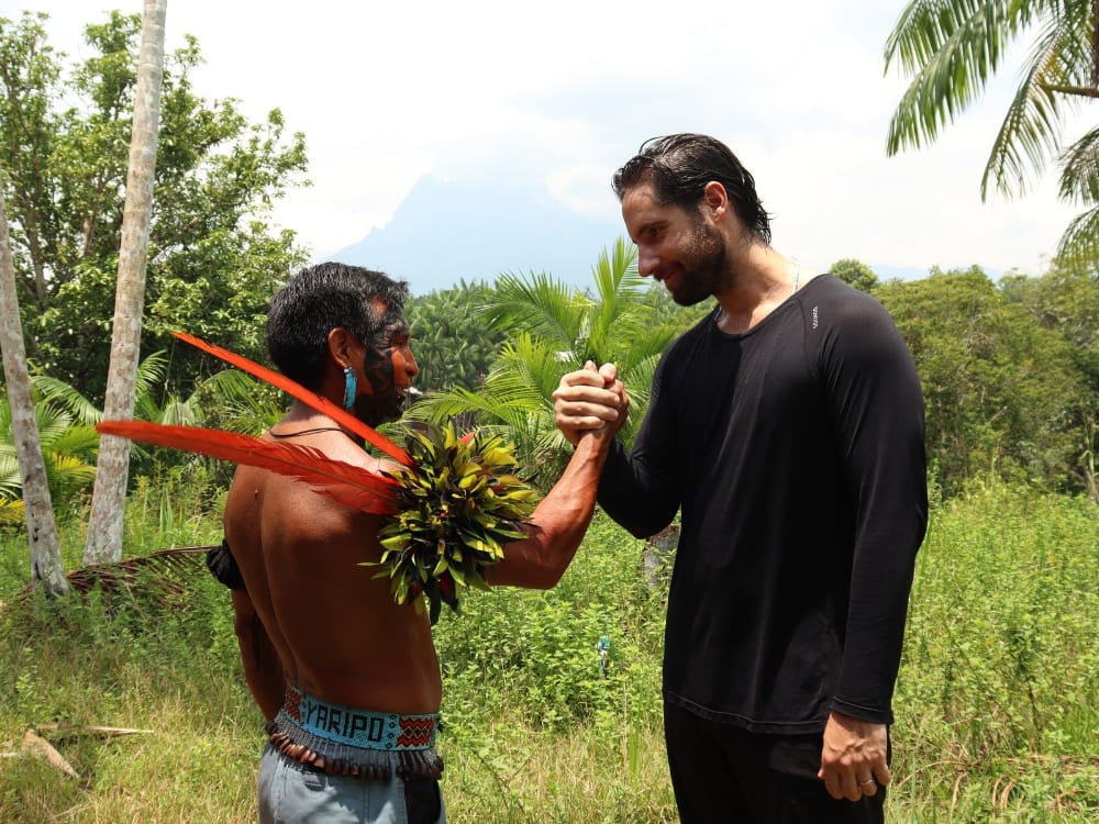Un guide autochtone Yanomami portant des plumes rouges traditionnelles serre la main d'un randonneur dans la forêt. Un guide autochtone Yanomami portant des plumes rouges traditionnelles serre la main d'un randonneur dans la forêt.